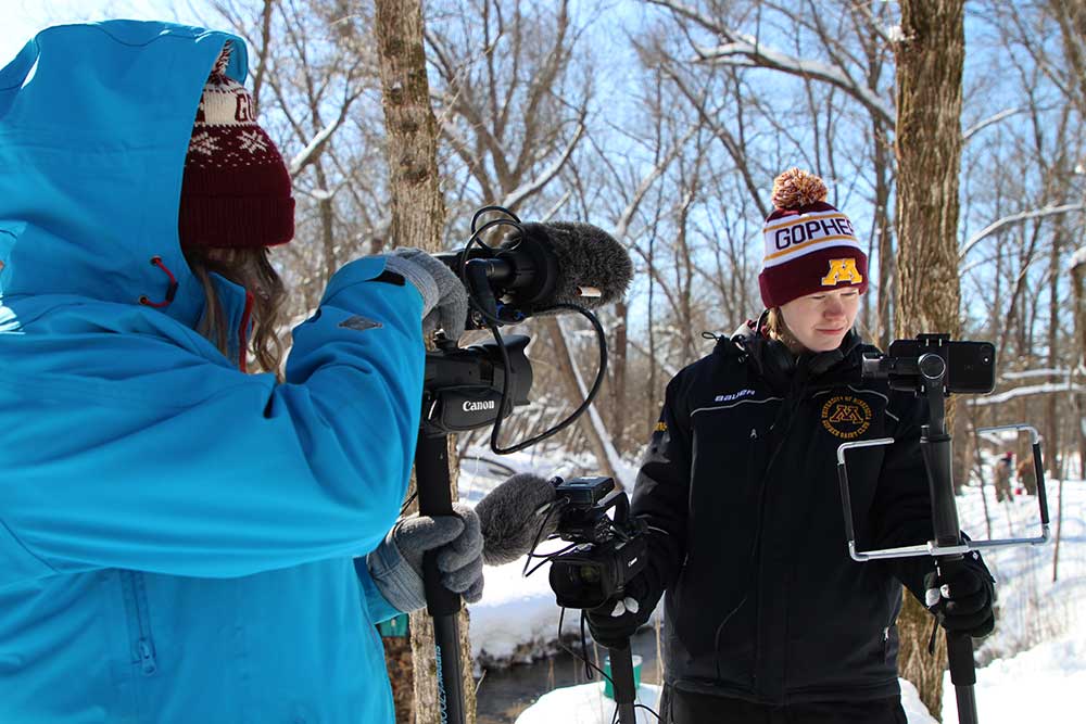 Two students work with cameras outside in winter among snow and bare trees.