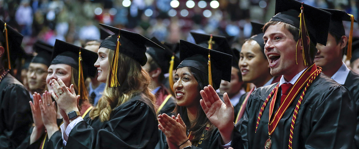 Wide shot of students in caps and gowns at graduation clapping and smiling