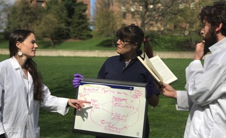 2 students in white lab coats describing some diagrams that are sketched on a dry erase board that is held by an instructor. 