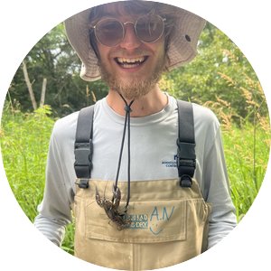 Avi Neumann smiles outside during his internship. A crayfish hangs on the string of his bucket hat.