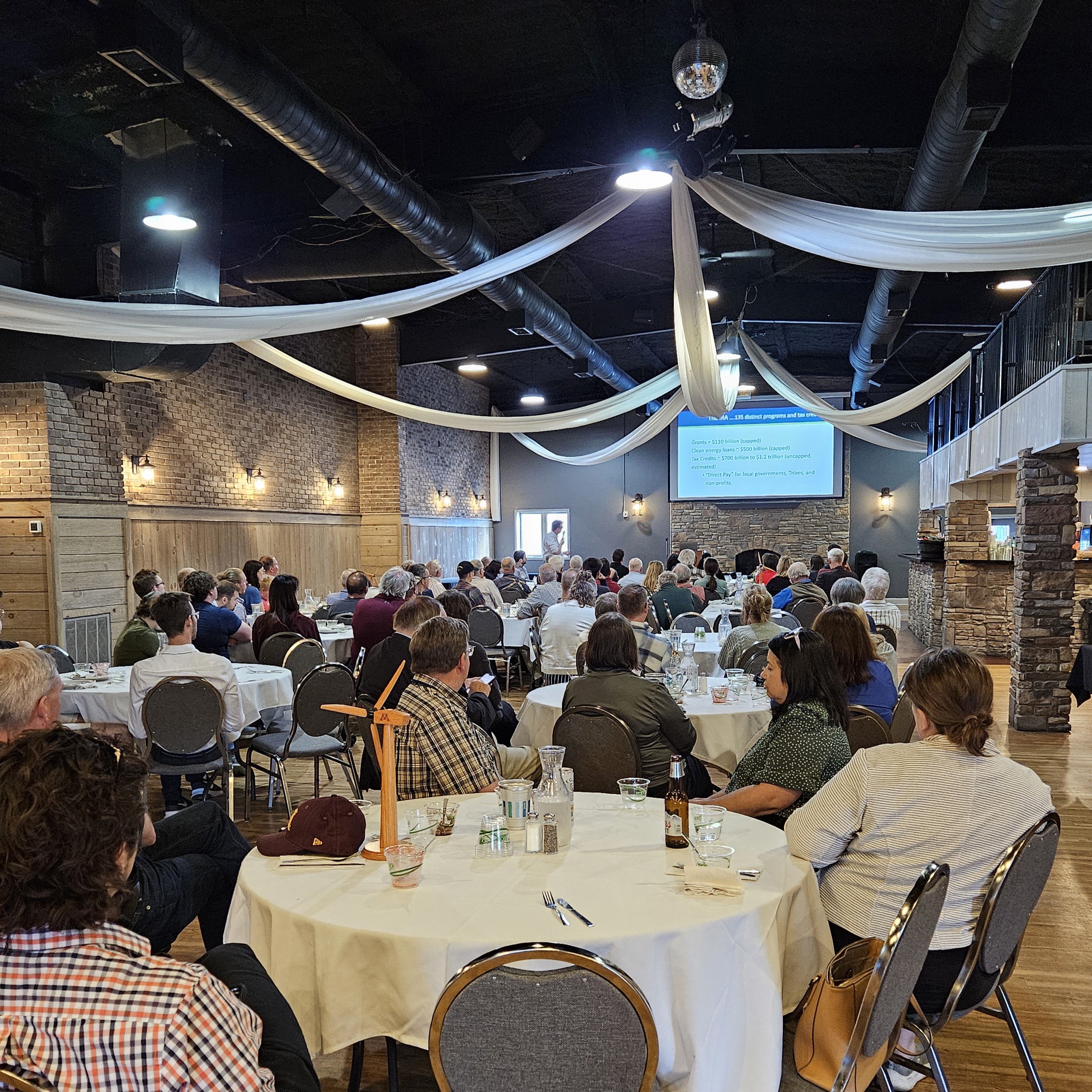 A large room is filled with people at circular tables, listening to a keynote presentation happening at the far end.