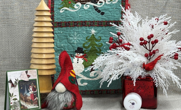 A variety of handmade items available for purchase at a previous auxiliary holiday sale event. Items include a felt decorative gnome, a seasonal greeting card, a quilt with winter motifs, a wooden pine tree, and a wintery floral arrangement. 
