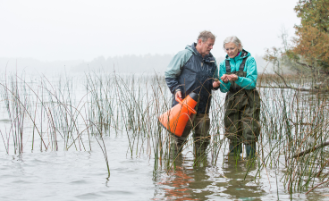 A man and a woman wade through a lake and examine an aquatic invasive species