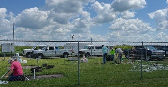 Several trucks parked around a fenced plot. In the foreground, MDA staff step around equipment on the ground as they install the new weather station.