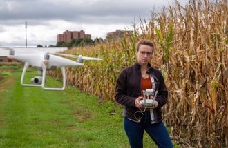 A woman stands next to a corn field and controls a white drone with a camera on it.