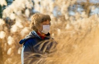 A woman in a blue coat and white face mask stand among tall golden grass.