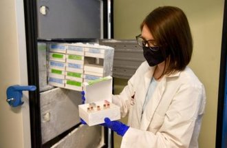 A woman with short brown hair, a black face mask and glasses takes samples out of a refrigerator in a lab.