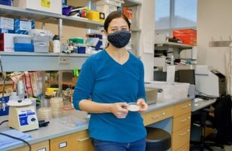 A woman wearing a blue shirt and a black face mask stands in a lab holding a petri dish.