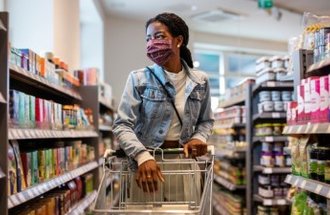 A woman in a jean jacket and a purple mask pushes a shopping cart down the aisle of a grocery store.