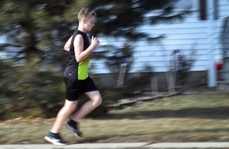 A boy in green and black jogs down the street on a sunny day.